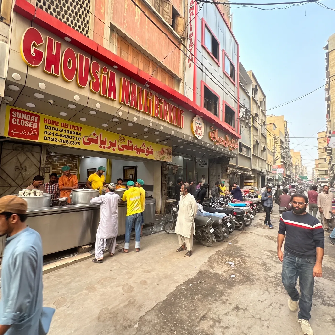 Street view of the Ghousia Nalli Biryani storefront in Liaquatabad, Karachi, with staff serving biryani from large steel pots at the counter and motorbikes parked along the busy street